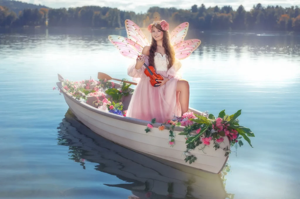 Fantasy-Fotoshooting Floral Boat am See in Solothurn, Model auf einem mit Blumen geschmückten Boot, trägt Feenflügel und hält eine Violine. Floral Boat Fotoevent on Elena Frizler Photography