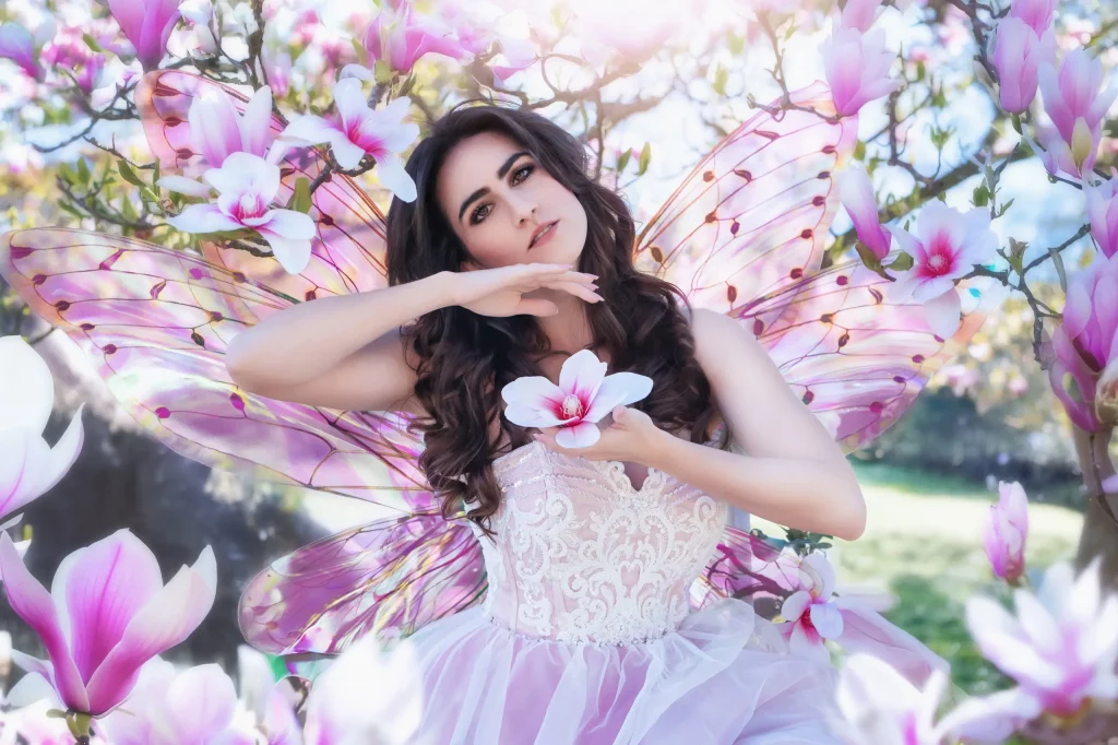 Model-im-rosa-Kleid-mit-Feenflügeln-in-Pastellfarben,-umgeben-von-Magnolienblüten,-hält-große-Blüte-in-der-Hand-–-Elena-Frizler-Photography