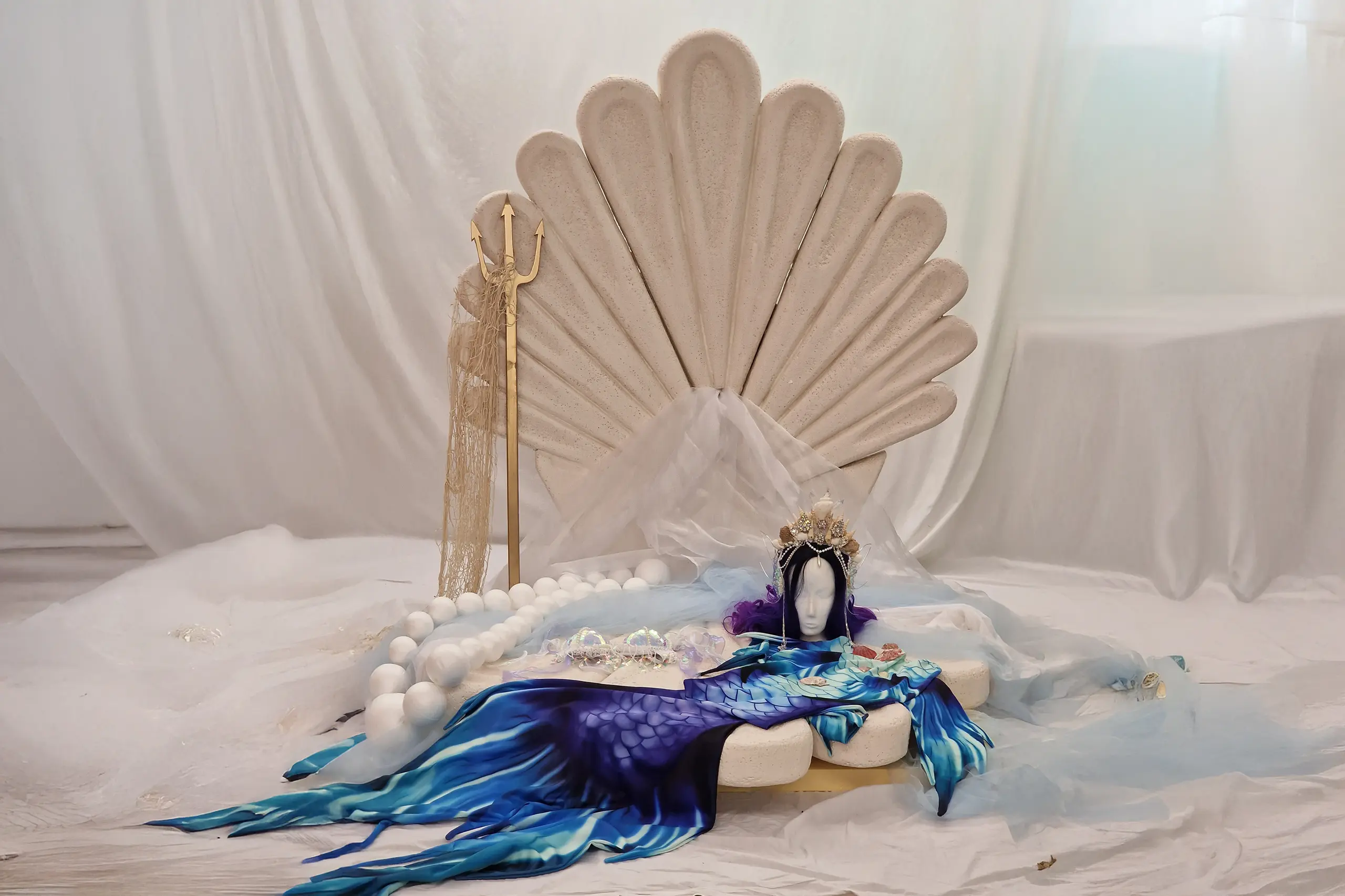 Große Fantasy-Muschel im Studio, dekoriert mit Tüllstoffen, Muscheln, Meerjungfrauenflosse und Dreizack vor weißem Hintergrund.
