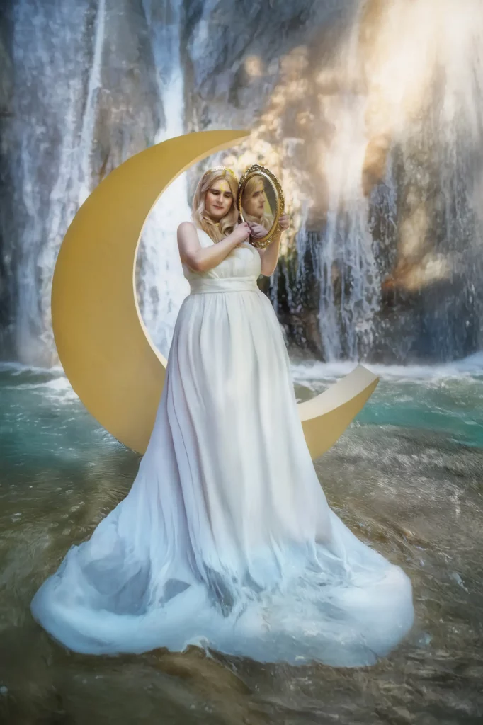 Stehendes Model vor Sichelmond, hält barocken Spiegel am Kopf, Gesicht spiegelt sich, Wasserfall im Hintergrund – Fantasy-Fotoshooting von Elena Frizler Photography, LuzernEntlebuch Schweiz.