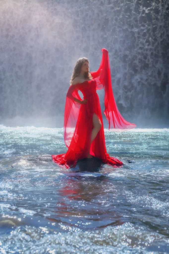 Model im roten Flying Dress steht vor einem gewaltigen Wasserfall, umgeben von Gischt und Licht – Fantasy Fotoshooting Schweiz von Elena Frizler Photography.