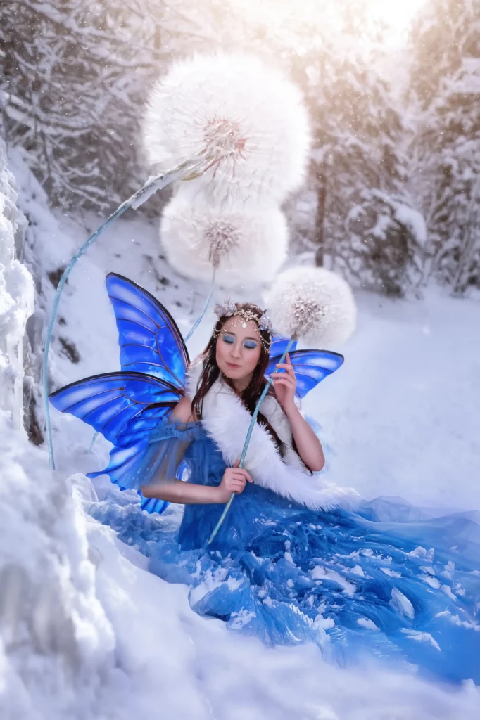 Fantasy Fairy Fotoshooting in Entlebuch Luzern Model im blauen Kleid mit Feenfluegeln im Schnee zwischen Pusteblumen
