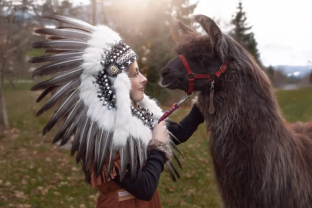 Indianer Fotoshooting Model mit grossem Kopfschmuck schaut vertraeumt einem Lama in die Augen