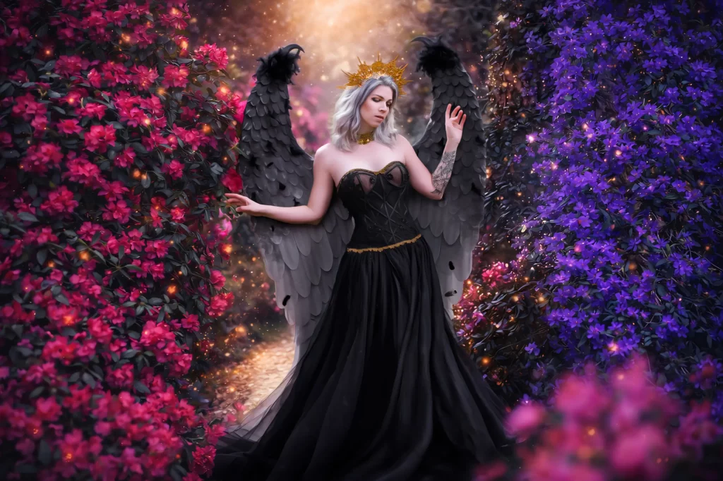 Model im schwarzen Kleid mit schwarzen Engelsflügeln steht auf kleinem Weg zwischen rosa und lila Rhododendronbüschen - Fantasy Dark Elena Frizler Photography