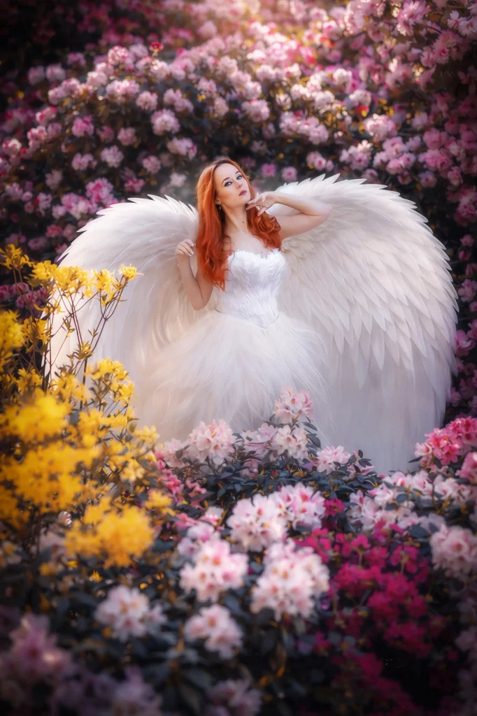 Model in weissem Kleid und grossen weissen Engelsfluegel posiert inmitten von Blumen in pastellfarben. Fotoshooting Fine Art von Elena Frizler Photograohy