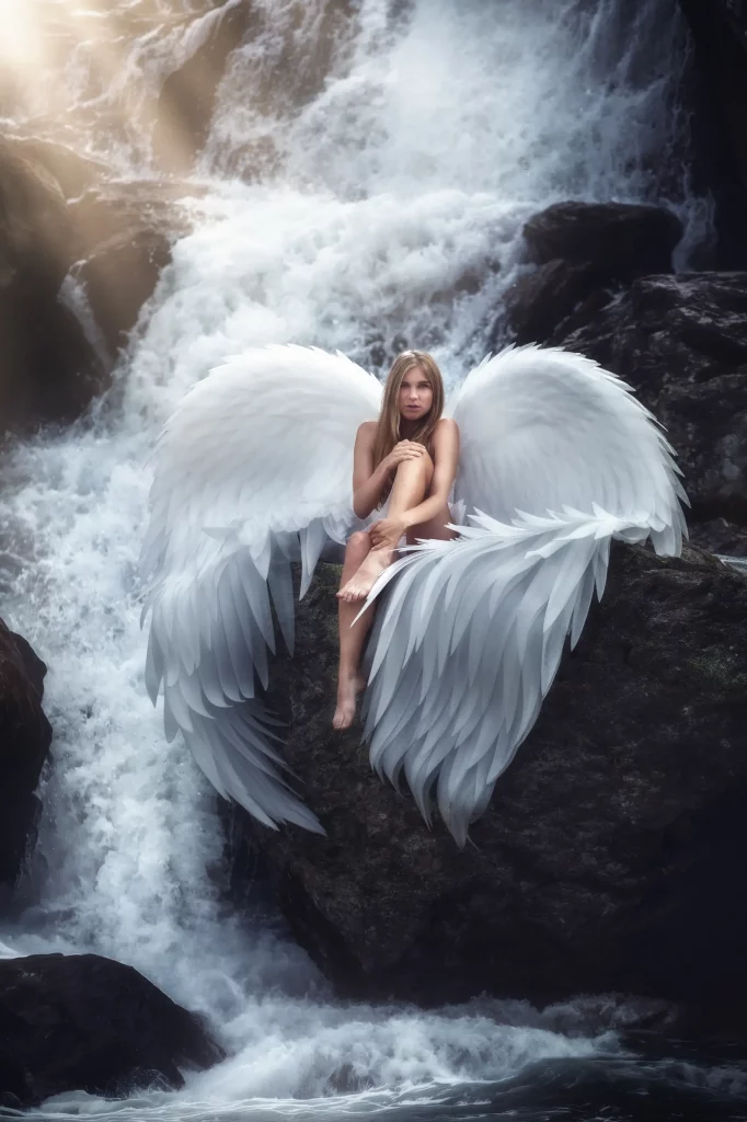 Engel mit grossen weissen Fluegeln auf einem Felsen vor einem Wasserfall waehrend eines Fantasy Fotoshootings in den Schweizer Alpen